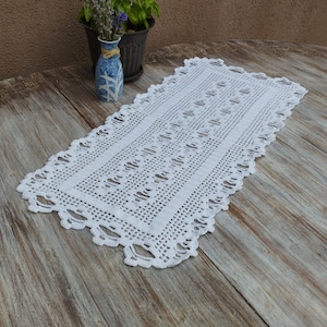 Può includere: Un runner da tavolo bianco all'uncinetto con un delicato motivo a pizzo. Il runner è disposto su un tavolo in legno rustico.