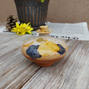 Op de afbeelding: Een kleine, ronde, keramische fles met een kurken stop. De fles is versierd met een handgeschilderd ontwerp van gele en blauwe vruchten.