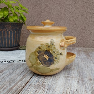 May include: A ceramic planter with a lid and two handles. The planter is decorated with a floral design in shades of green, brown, and black. The planter is sitting on a wooden surface.