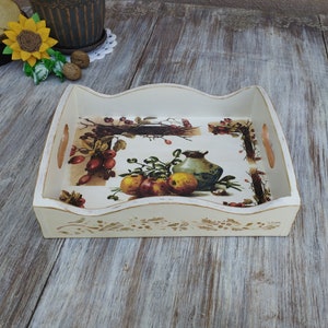 Könnte beinhalten: Weißes Holztablett mit einem dekorativen Blumendesign und einem Vintage-Druck von Obst und einer Kanne. Das Tablett hat einen gewellten Rand und zwei Griffe.