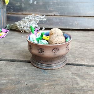 Pode incluir: Uma tigela de cerâmica marrom com um design decorativo, cheia de doces e nozes variados. A tigela tem uma borda larga e uma base arredondada.