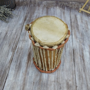 May include: A handmade drum with a light tan drumhead and a cylindrical body. The body is constructed from vertical, light-coloured strips, bound by reddish-brown trim at the top and bottom. The drum is likely made of natural materials.