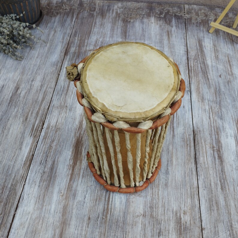 May include: A handmade drum with a light tan drumhead and a cylindrical body. The body is constructed from vertical, light-coloured strips, bound by reddish-brown trim at the top and bottom. The drum is likely made of natural materials.