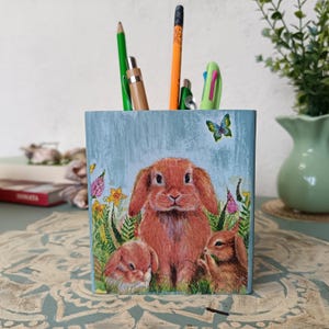 May include: A blue wooden pencil holder with a painted design of three bunnies in a field of flowers. The bunnies are brown and white with pink noses. There are butterflies and green leaves in the background.