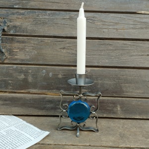 Pu&ograve; includere: Un portacandele in metallo caratterizzato da una cornice quadrata con volute decorative e un inserto centrale in vetro blu circolare. Il supporto presenta una piccola coppa superiore per una candela affusolata bianca su base piatta.