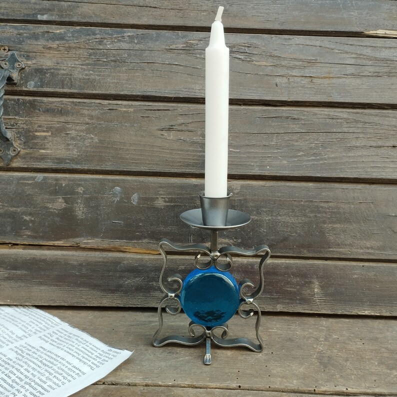 Pu&ograve; includere: Un portacandele in metallo caratterizzato da una cornice quadrata con volute decorative e un inserto centrale in vetro blu circolare. Il supporto presenta una piccola coppa superiore per una candela affusolata bianca su base piatta.