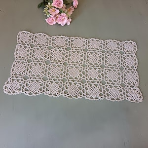 May include: Cream-coloured, rectangular crochet doily with a repeating geometric pattern. The edges are scalloped. A small bouquet of pink roses sits in the upper left corner. The doily is displayed on a muted green surface.