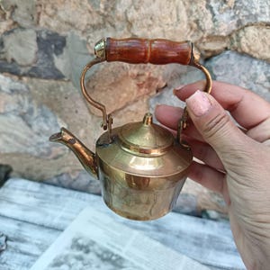 May include: A miniature brass teapot featuring a polished metallic finish, a conical lid, and a curved spout. The teapot includes a hinged metal handle topped with a smooth, cylindrical brown wooden grip for holding the small vessel.