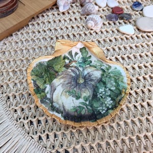 Op de afbeelding: Decoratieve zeeschelp met een pompoen- en bladerenontwerp. De afbeelding toont een grote, witte en grijze pompoen omringd door groene bladeren en witte bloemen. De schelp heeft een goudkleurige rand.