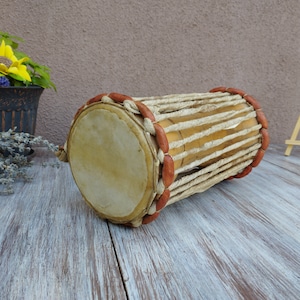 May include: A handcrafted, cylindrical drum with a light brown wooden body and tan leather drumheads. The drum is wrapped with natural-coloured rope and accented with reddish-brown beads. The drum is lying on a weathered, light-coloured wooden surface.