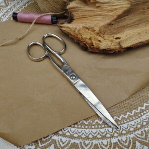 May include: Silver scissors with a black handle on a brown paper background. The scissors have the word "D.L.L" printed on the blade.