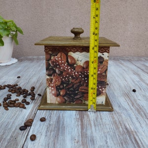 May include: A square wooden box with a hinged lid and a brown knob. The box is decorated with a collage of coffee beans, brown ribbon, and musical notes. The box is approximately 5 inches tall.