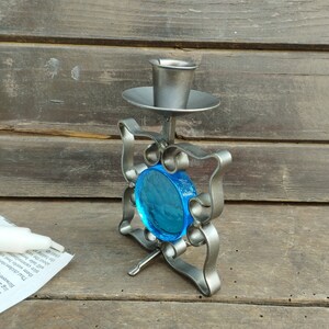 Pu&ograve; includere: Un portacandele in metallo con cornice quadrata, volute decorative e un inserto centrale in vetro blu circolare. L'oggetto presenta una coppa superiore per una candela e un piccolo piedino di supporto in metallo alla base.