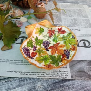 Może przedstawiać: Ozdobna muszla z pozłacaną krawędzią, ozdobiona wzorem decoupage z winogron i jesiennych liści w odcieniach fioletu, zieleni i pomarańczy. Muszla leży na gazecie.
