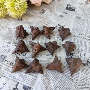 May include: A collection of eleven dried, dark brown water caltrop seed pods, each featuring a distinct four-pointed, horned shape with a central circular opening, arranged in a loose cluster on a surface printed with vintage newspaper text.