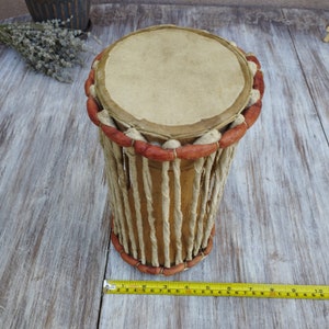 May include: A cylindrical drum with a light brown leather drumhead and a wooden body. The drum's body is wrapped with twisted rope and accented with red-brown wooden beads. The drum is approximately 25 cm long.