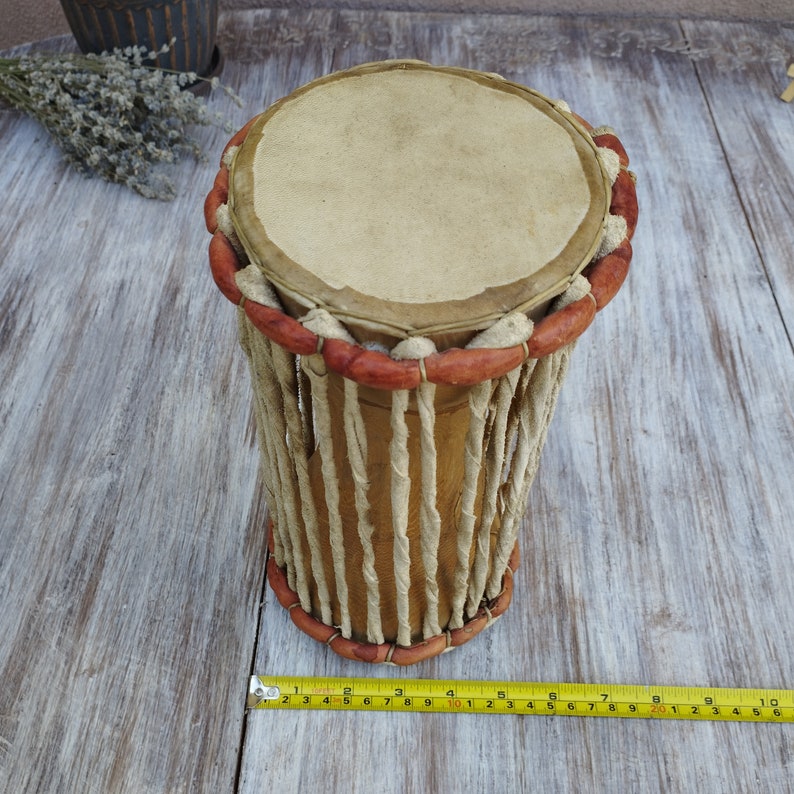 May include: A cylindrical drum with a light brown leather drumhead and a wooden body. The drum's body is wrapped with twisted rope and accented with red-brown wooden beads. The drum is approximately 25 cm long.