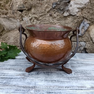 May include: A hammered copper pot with a dark brown metal stand. The pot has a rounded body and two curved handles. The stand has three feet. The pot's interior has a green patina.