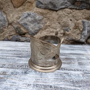 May include: A silver-colored, vintage teacup holder with an ornate, engraved design. The holder features a circular base, a decorative band, and a handle. The design includes floral and scroll patterns. The background is a weathered stone wall.