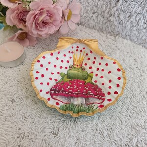 May include: A white seashell with a gold rim, painted with red polka dots and a green frog wearing a gold crown sitting on a red mushroom.