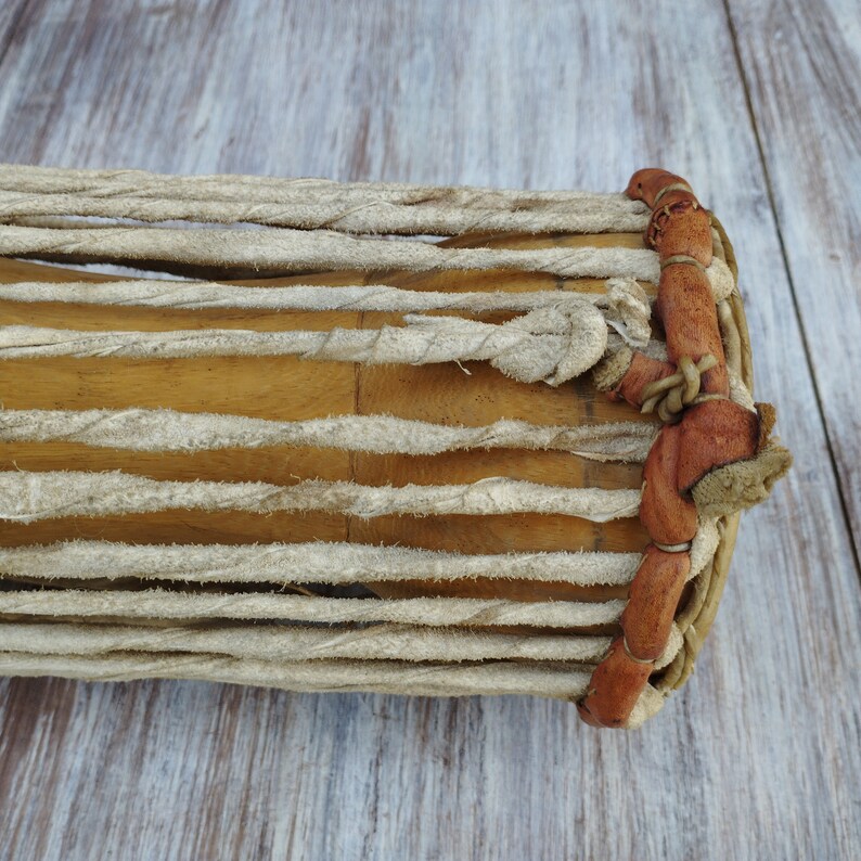 May include: Close-up of a wooden drum with a light brown finish. The drum is wrapped with thick, off-white rope and has brown leather accents on the side. A percussion instrument.