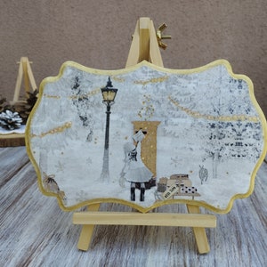 May include: A white and gold wooden sign with a winter scene. The sign features a girl walking towards a mailbox with a street lamp in the background. The sign is on a small wooden easel.