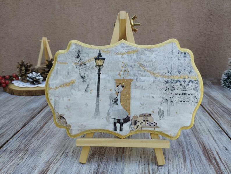 May include: A white and gold wooden sign with a winter scene. The sign features a girl walking towards a mailbox with a street lamp in the background. The sign is on a small wooden easel.