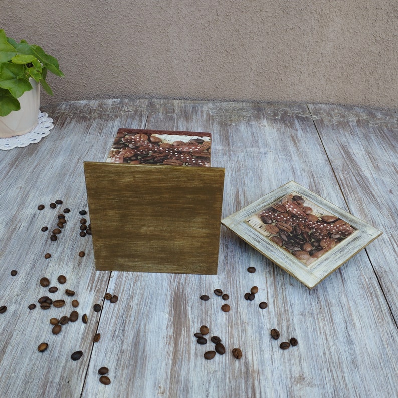 May include: A wooden box with a brown and white design featuring coffee beans. The box is open and the inside is visible. The box is sitting on a white wooden surface with coffee beans scattered around it.