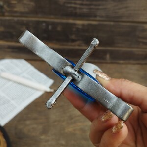 Pu&ograve; includere: Uno spegnicandela in metallo forgiato a mano con un manico lungo e piatto e una parte superiore a forma di croce. Il metallo presenta una finitura industriale scura e ruvida con punti di saldatura visibili al centro della struttura.