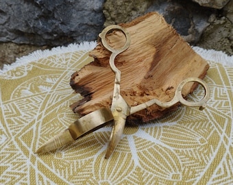 May include: Brass antique scissors with a ring handle, resting on a piece of wood and a patterned fabric.