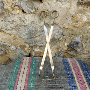 May include: A pair of silver-toned metal tongs with a crossed design. The tongs are on a striped fabric background.