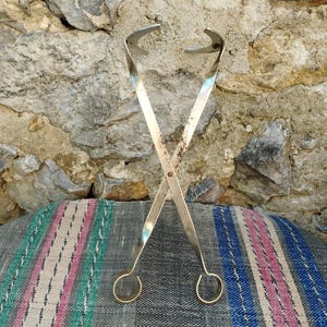May include: A pair of silver-toned metal tongs with a loop at each end. The tongs are crossed and resting on a striped fabric surface.