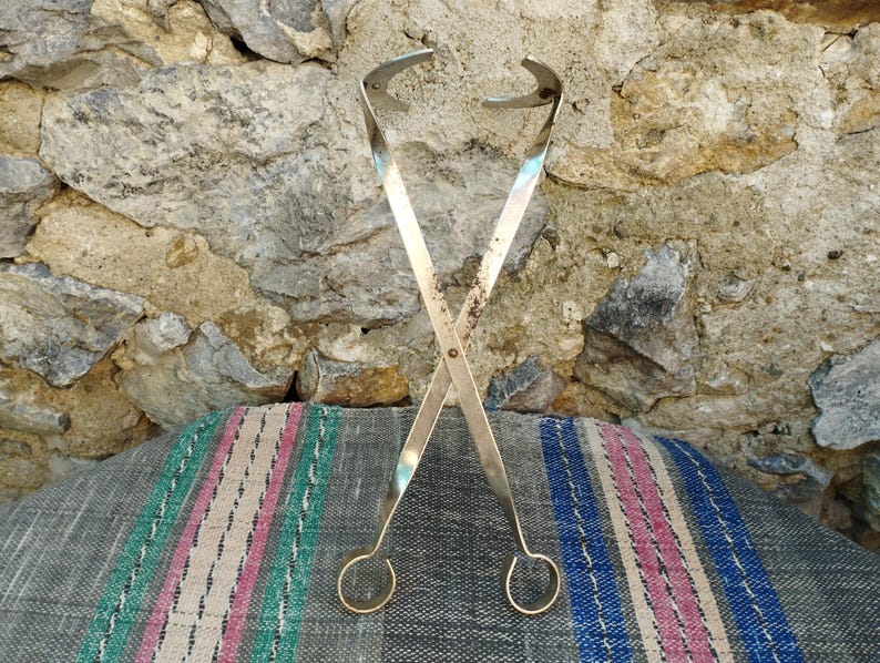 May include: A pair of silver-toned metal tongs with a loop at each end. The tongs are crossed and resting on a striped fabric surface.