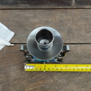 Pu&ograve; includere: Un portacandele grigio metallizzato con una parte superiore circolare e accenti laterali decorativi, posizionato accanto a un metro a nastro giallo che mostra una larghezza di circa 10 cm. Il portacandele presenta un'apertura centrale.