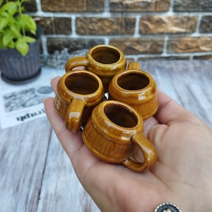Puede incluir: Cuatro peque&ntilde;as tazas de cer&aacute;mica marr&oacute;n con asas. Las tazas est&aacute;n dispuestas en c&iacute;rculo y est&aacute;n siendo sostenidas en la mano de una persona.