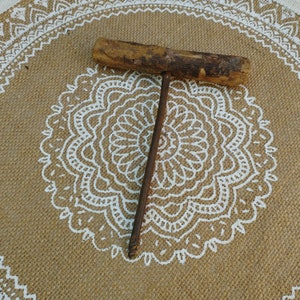 May include: A rusty metal hand drill with a wooden handle. The drill is lying on a brown burlap surface with a white mandala design.