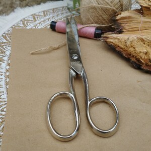May include: A pair of silver-toned scissors with a black handle on a brown surface. The scissors have a small inscription that reads "PLY."