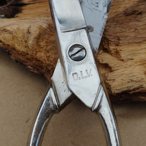 May include: Close-up of a silver pair of scissors with a black handle. The scissors have a  "O.L.V." marking on the blade.