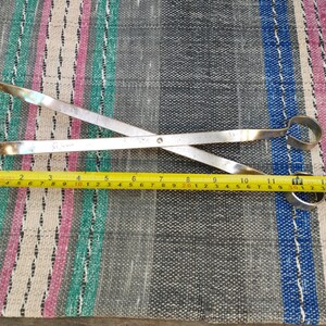 May include: A pair of silver metal tongs with a measurement tape showing 15 inches in length. The tongs are crossed and have a circular opening at each end.