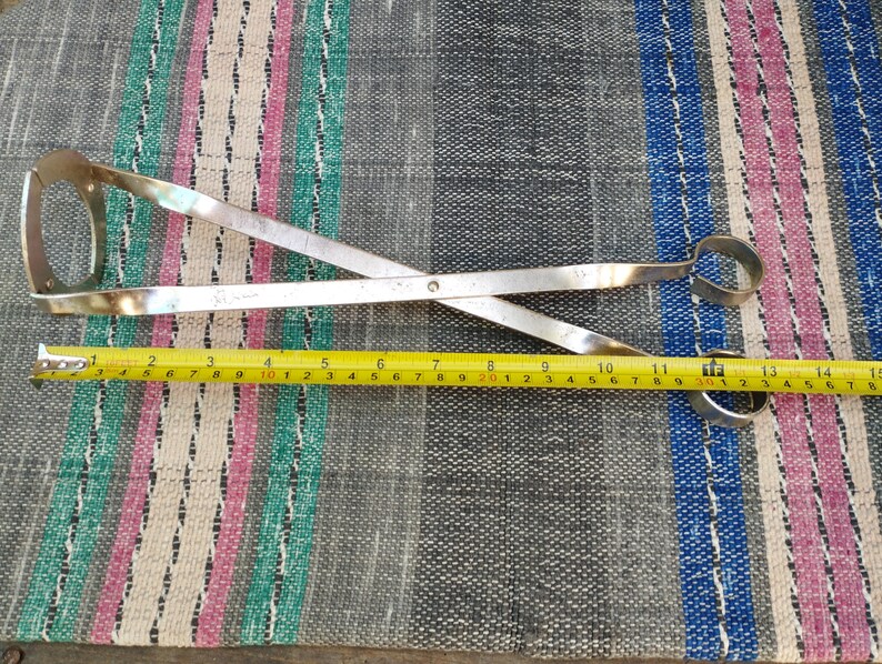 May include: A pair of silver metal tongs with a measurement tape showing 15 inches in length. The tongs are crossed and have a circular opening at each end.