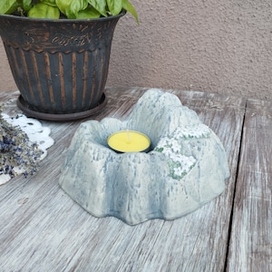 Könnte beinhalten: Ein grauer, bergförmiger Kerzenhalter mit einer gelben Kerze im Inneren. Der Halter hat eine strukturierte Oberfläche und ein kleines Blumendesign. Eine Topfpflanze befindet sich im Hintergrund.