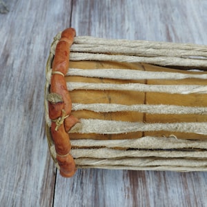 May include: Close-up of a cylindrical drum with a light brown wooden body and tan leather accents. The drum's body is made of parallel wooden slats, bound together with tan leather strips and rope. The ends are wrapped in brown leather.