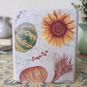 Puede incluir: Una caja de madera blanca con un dise&ntilde;o de oto&ntilde;o. La caja presenta un girasol, dos calabazas y bayas rojas. El dise&ntilde;o est&aacute; impreso en la caja y tiene un aspecto vintage.