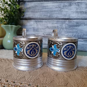 May include: Two silver metal cups with blue and white floral designs on a brown background. The cups have a handle and are made of metal.