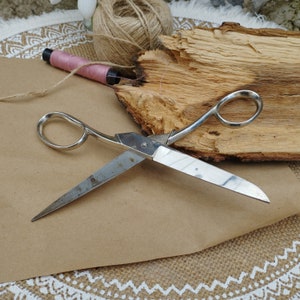 May include: A silver pair of scissors with a wooden background. The scissors have a single blade and a rounded handle.