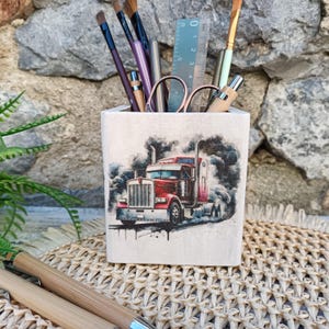 Könnte beinhalten: Ein quadratischer, cremefarbener Stifthalter mit einem Aquarellbild eines roten Sattelschleppers, der schwarzen Rauch ausstößt. Der Halter enthält verschiedene Kunstutensilien, darunter Pinsel, Scheren und ein Lineal. Der Stifthalter steht auf einer gewebten Matte.
