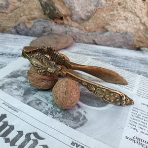 Può includere: Uno schiaccianoci antico in bronzo con dettagli floreali ornati, appoggiato su un giornale con due noci. Le maniglie dello schiaccianoci sono curve e le ganasce sono aperte, pronte a rompere le noci. Lo sfondo presenta un muro di pietra.