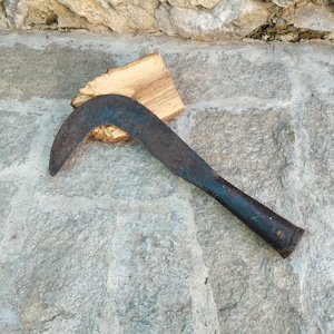 May include: A rusty, curved, black metal hand tool with a wooden handle. The tool is lying on a gray stone surface.