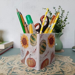 Puede incluir: Un porta l&aacute;pices de cer&aacute;mica blanca con un dise&ntilde;o de oto&ntilde;o con girasoles, calabazas y sand&iacute;as. El porta l&aacute;pices est&aacute; lleno de l&aacute;pices, bol&iacute;grafos y tijeras.