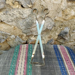 May include: A pair of silver-toned metal tongs with a crossed design. The tongs are on a striped fabric background.